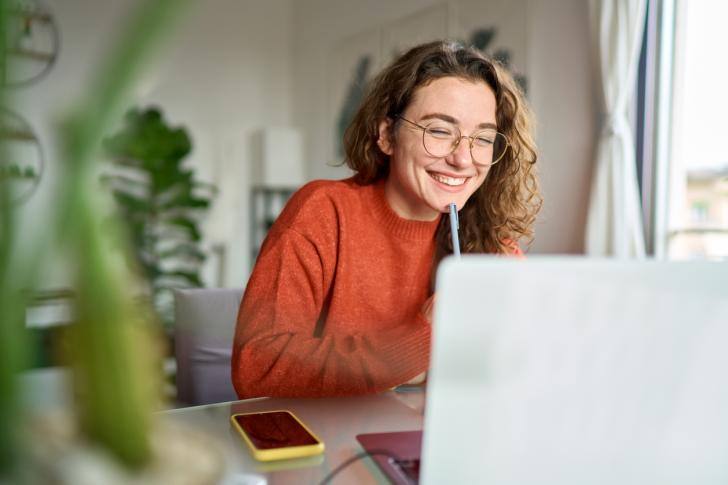 A woman in an orange sweater smiles at her laptop in a bright room with plants, perhaps exploring her educational journey in natural sciences at Bastyr University.