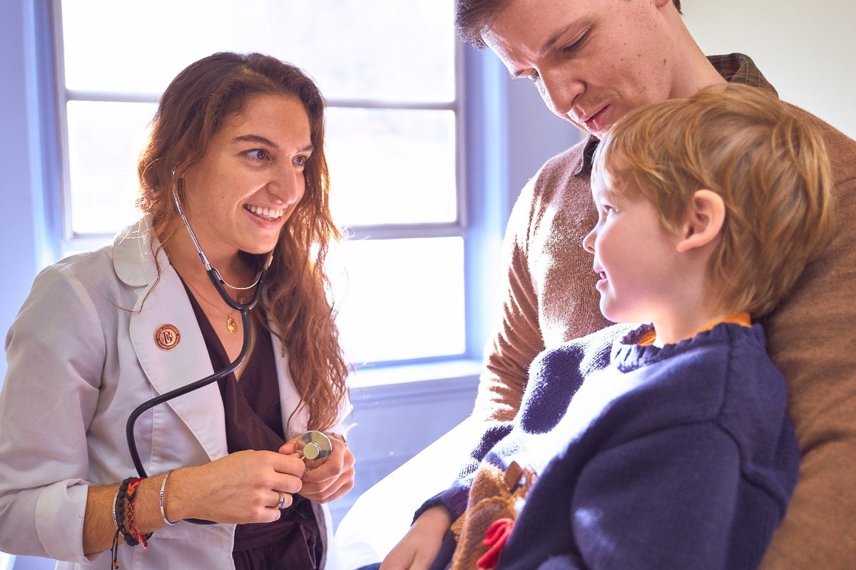 A doctor in a white coat smiles while interacting with a young boy sitting on a man's lap in a brightly lit room. A doctor in a white coat smiles while interacting with a young boy sitting on a man's lap in a brightly lit room.